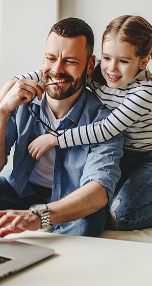Researching an Allegiance Auto Loan is easy for this dad on his laptop while his daughter hugs him.